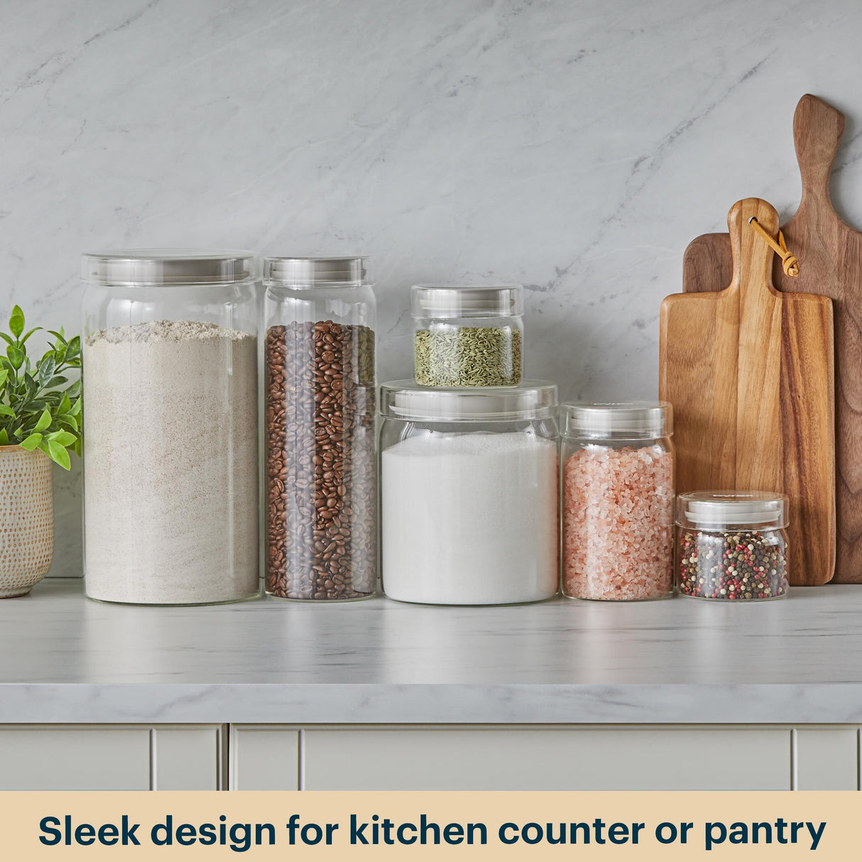 Glass jars with lids on a kitchen counter with wooden cutting boards and a plant, against a light gray marble wall.