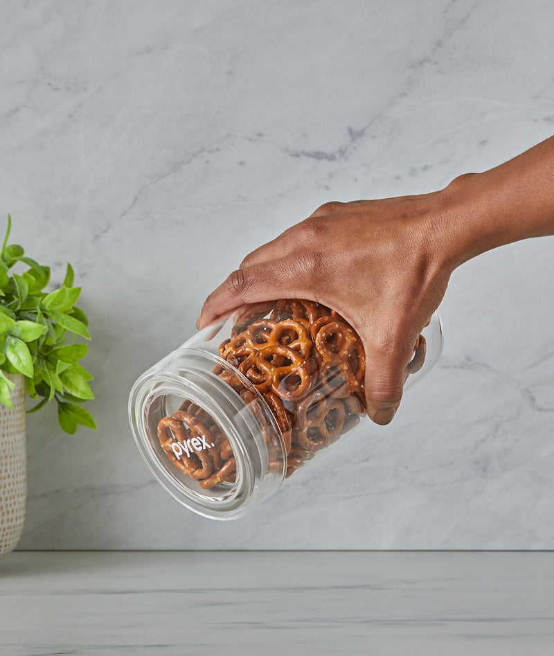 Hand holding a Pyrex jar filled with pretzels on a marble surface.