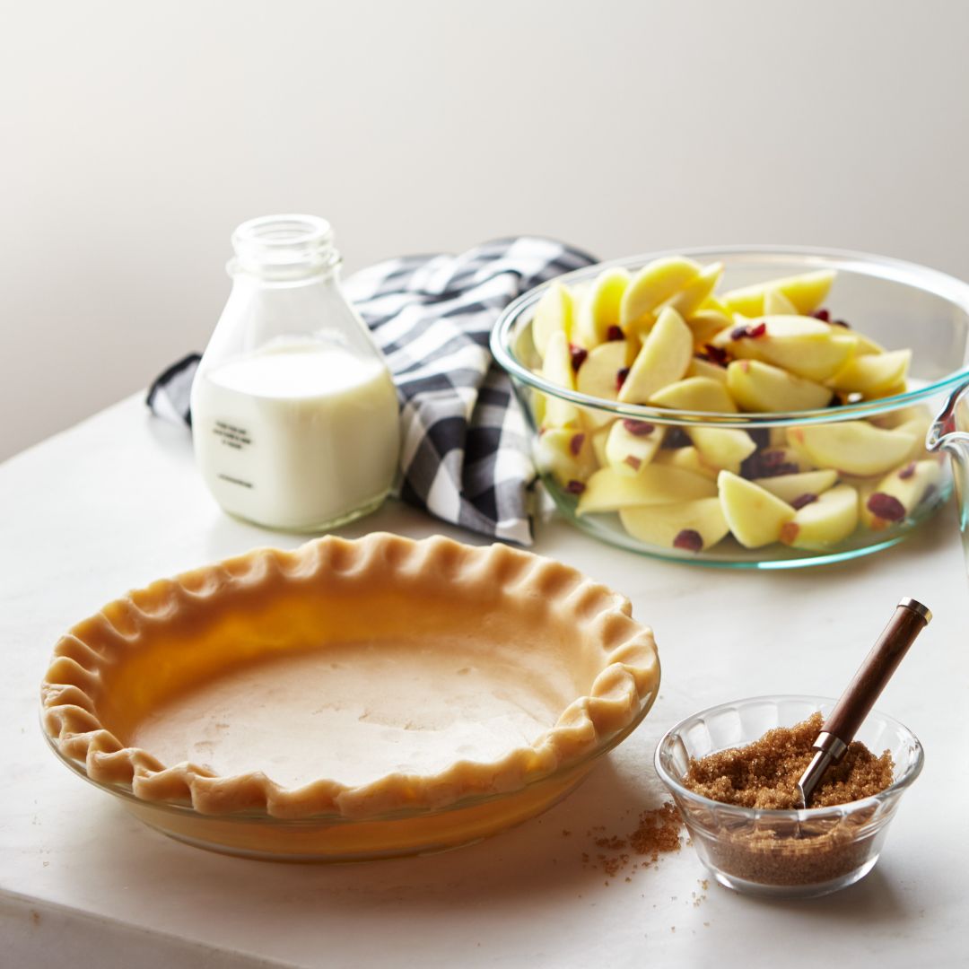 Pie plate with dough and brown sugar next to it with apples and milk on the background