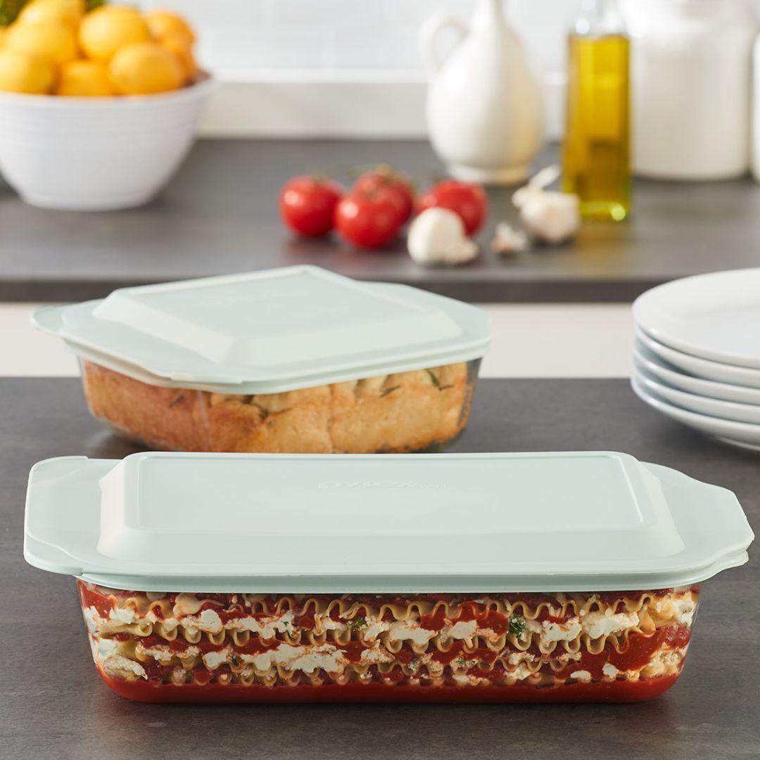 Two glass lasagna dishes with lids on a kitchen counter with ingredients in the background.